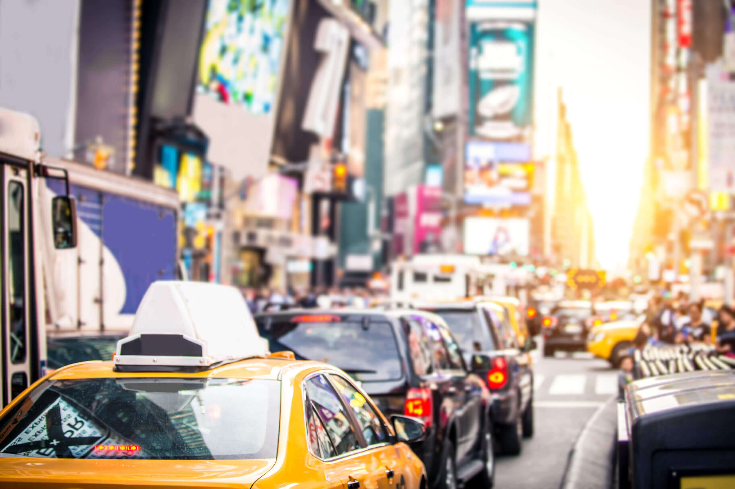 Traffic jam with yellow taxis and other cars on a busy city street, likely Times Square in New York City, with bright billboards and buildings in the background and sunlight shining ahead.