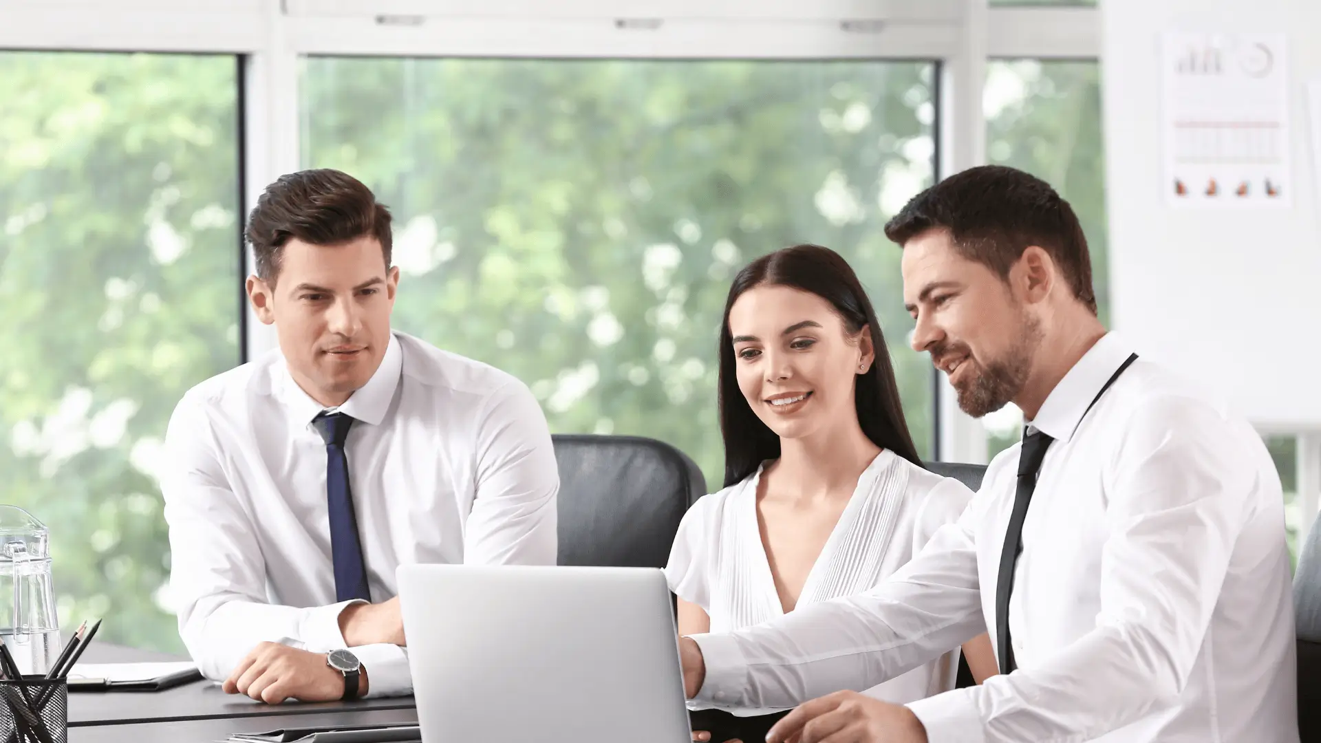 Three business professionals, two men and one woman, sit at a desk in an office, looking at a laptop screen and smiling. Large windows with greenery outside are in the background.
