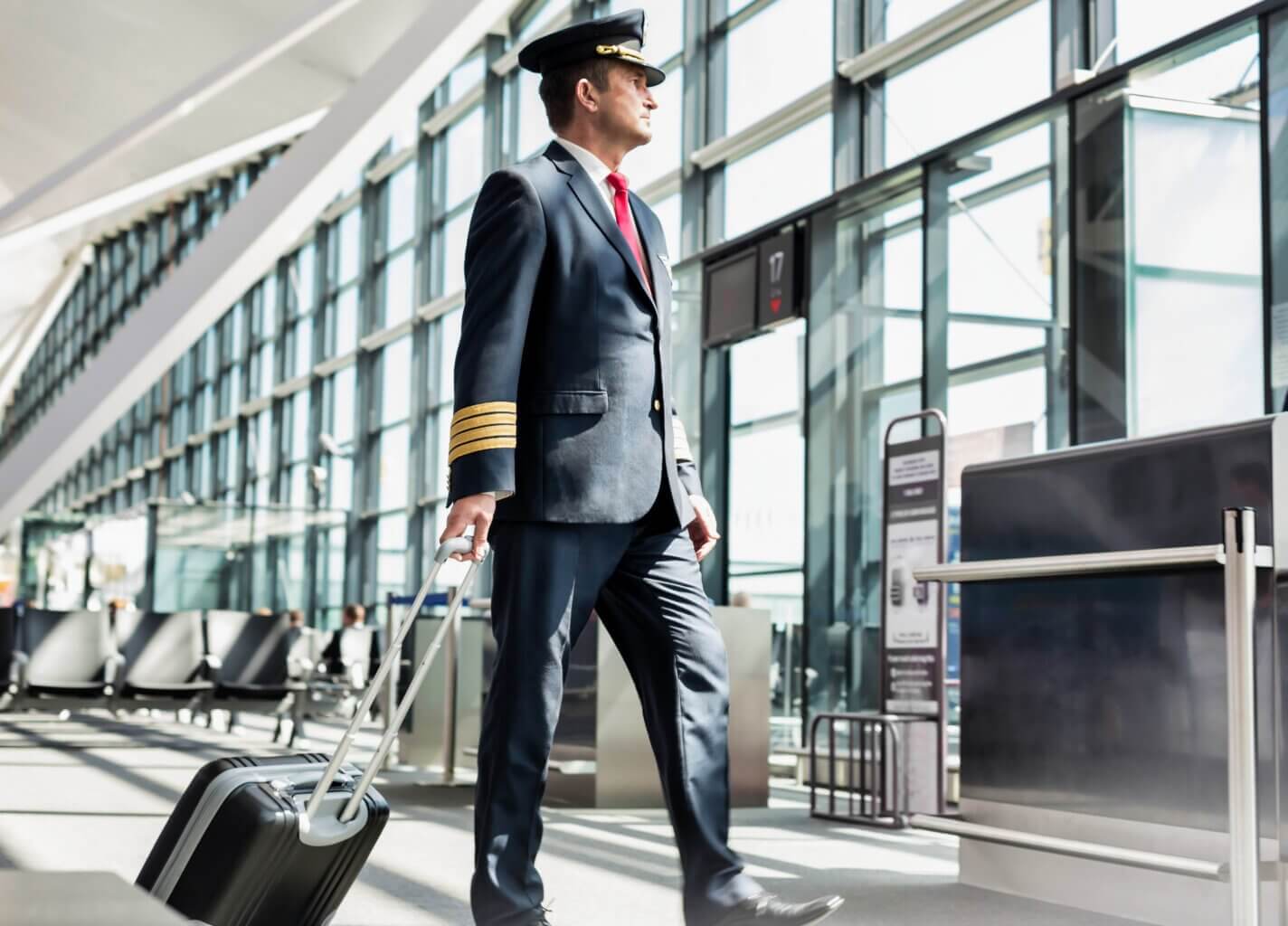 A commercial airline pilot in uniform and cap walks through a bright airport terminal, pulling a rolling suitcase, with empty seats and large windows in the background.