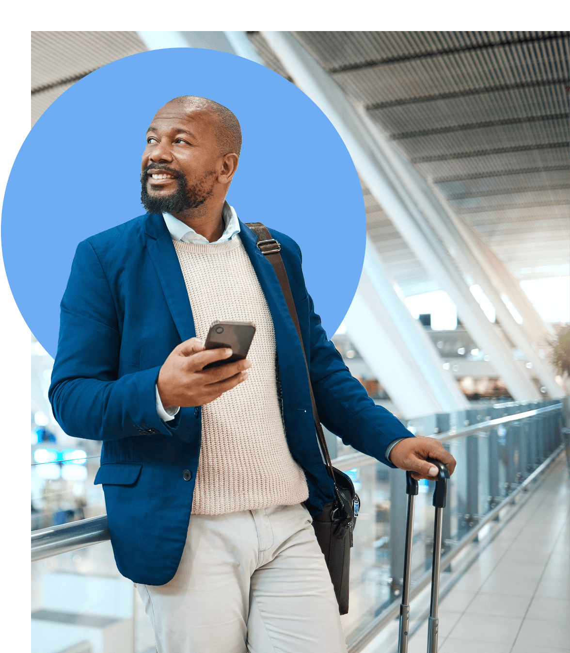 A man in a blue blazer and light sweater smiles while standing in an airport, holding a smartphone and suitcase, with a crossbody bag over his shoulder. A large blue circle is behind him.