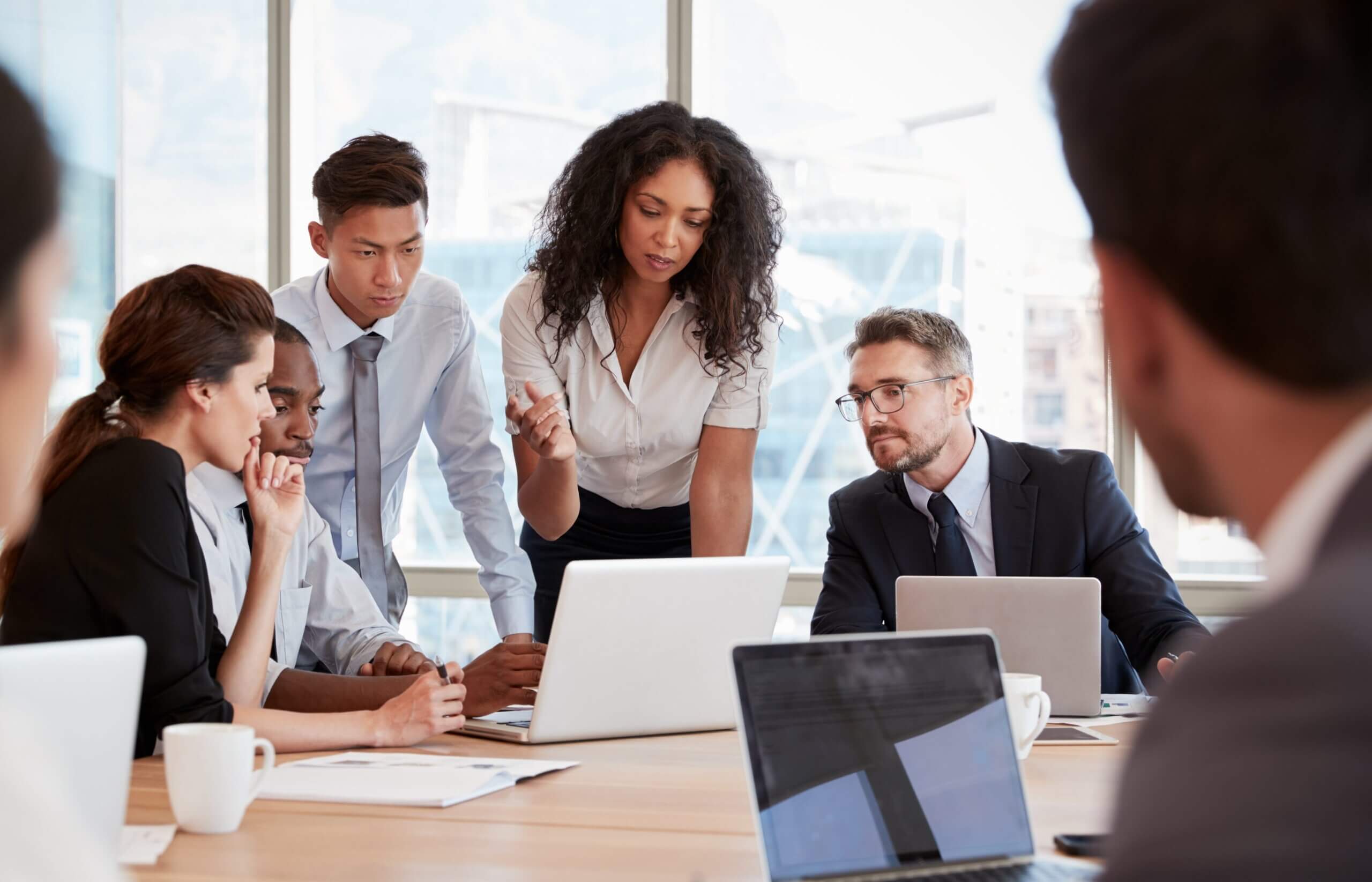 A group of business professionals in formal attire are gathered around a table in an office, focused on a laptop as one woman gestures while speaking, suggesting a collaborative discussion or meeting.