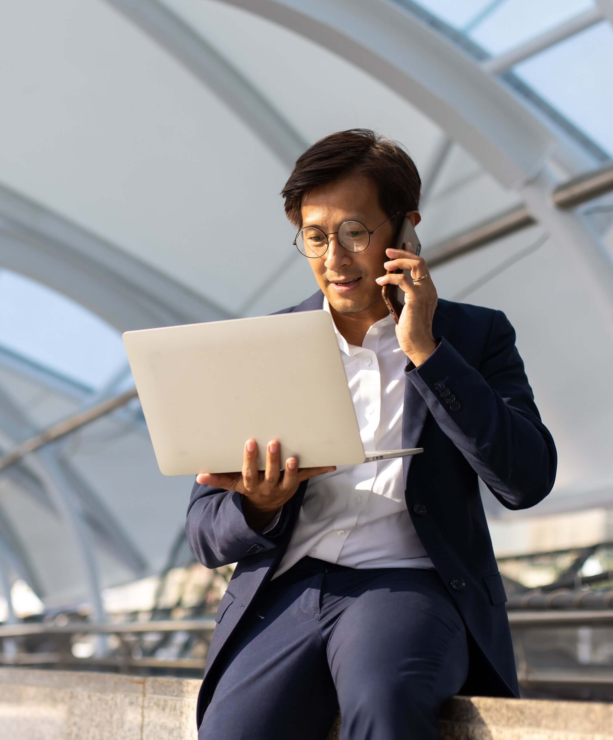 A person in a suit sits outdoors, holding a laptop and talking on a smartphone, with a modern architectural structure in the background—perfectly embodying the fast-paced environment of HK Company Formation.
