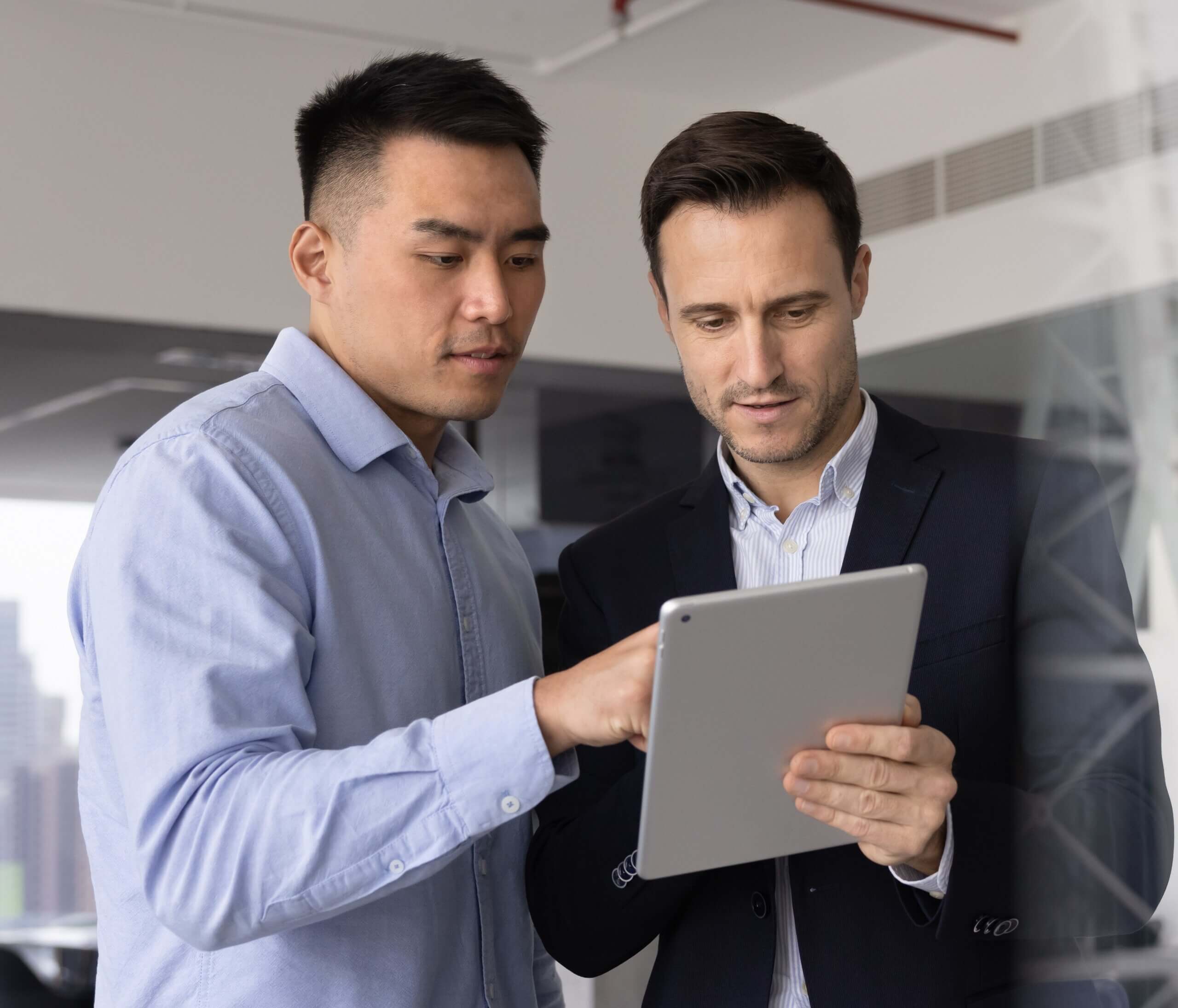 Two men standing in an office, looking at and discussing content on a tablet. One is wearing a light blue shirt, and the other is in a dark suit. They appear to be collaborating on work, possibly reviewing HK Due Diligence Searches together.