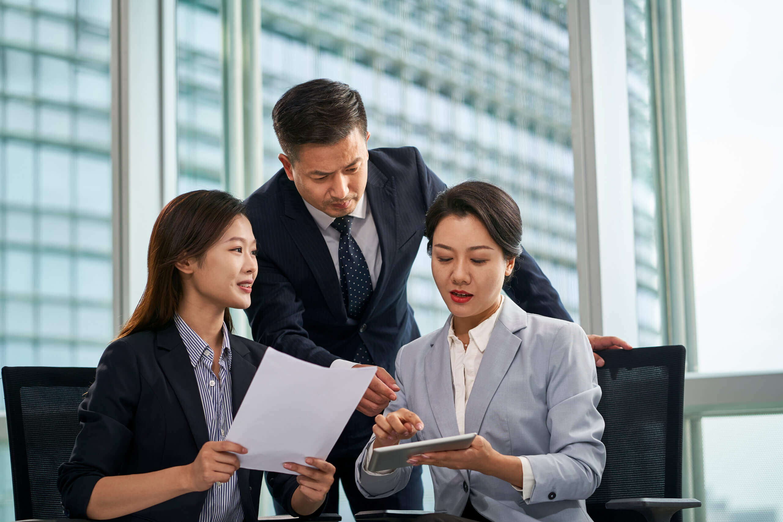 Three business professionals in formal attire discuss documents and a tablet device in a modern office with large windows and a cityscape background.