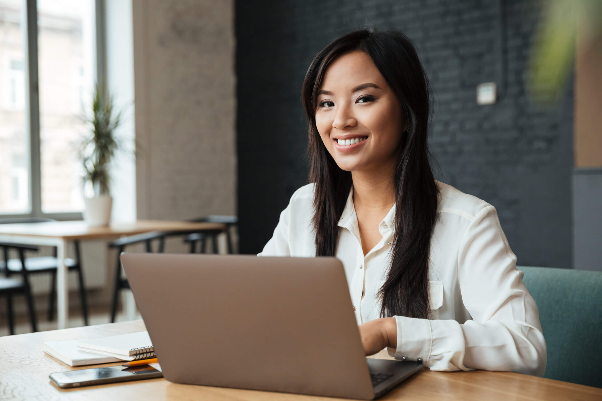 A woman with long dark hair, wearing a white blouse, sits at a desk smiling while working on a laptop. Notebooks, a pen, and a phone are on the desk. The background shows a modern, bright office.