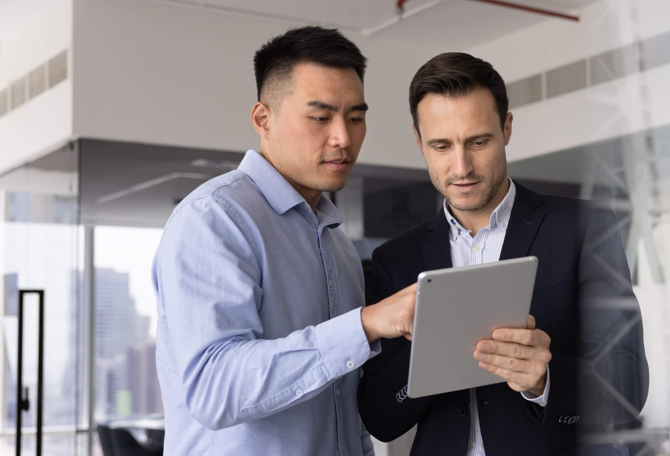 Two men in business attire stand in an office, looking at and discussing something on a tablet device. One man gestures towards the screen while the other listens attentively.