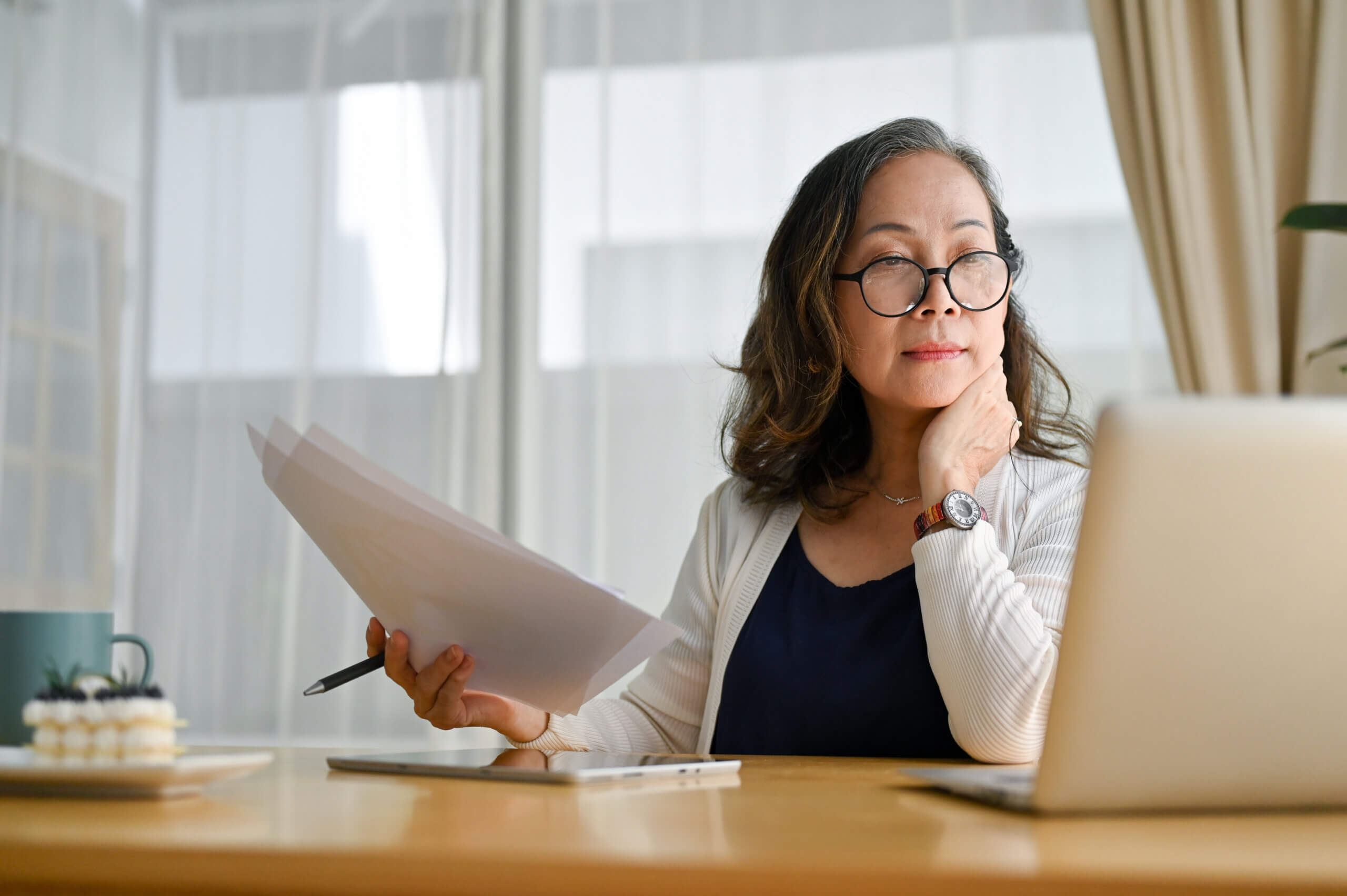A woman with glasses sits at a desk, holding papers and a pen, looking thoughtfully at a laptop. There is a coffee cup and a dessert on the table. Light streams in through sheer curtains behind her.