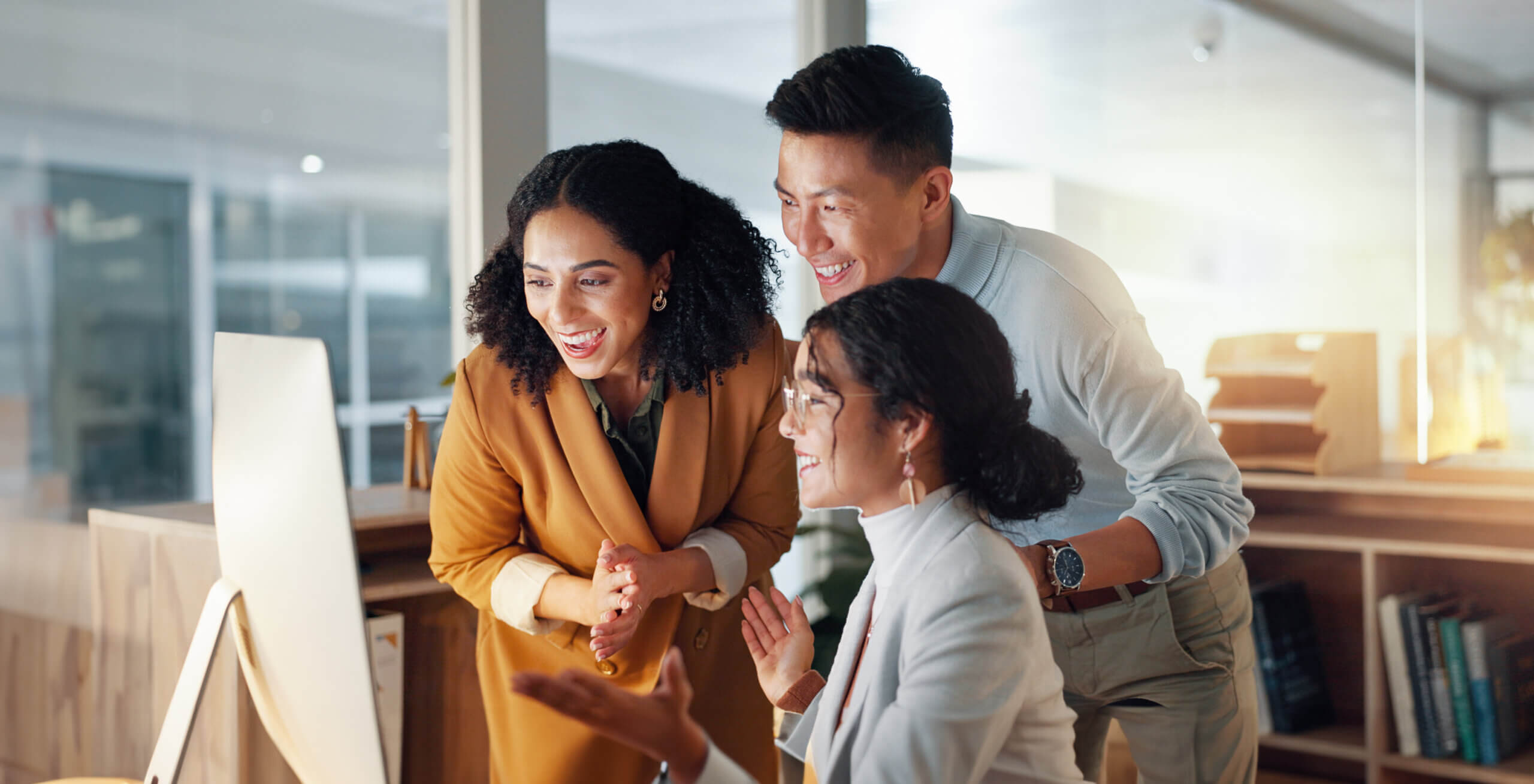 Three people in business attire smile and look at a computer screen together in a modern office, appearing engaged and happy, with natural light illuminating the workplace.