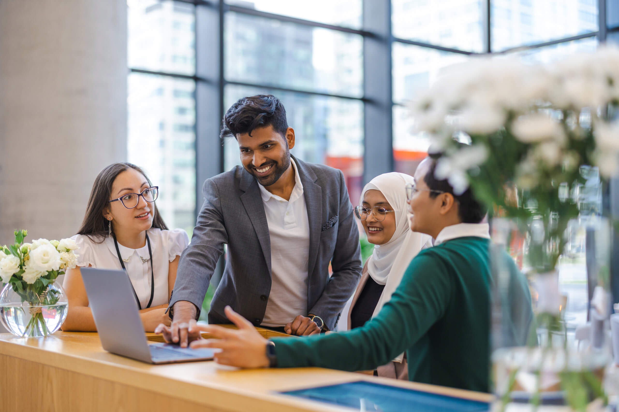 Four people gather around a laptop at a modern office desk, engaged in discussion. One person is pointing at the laptop screen while others listen and smile. Large windows and flowers are in the background.