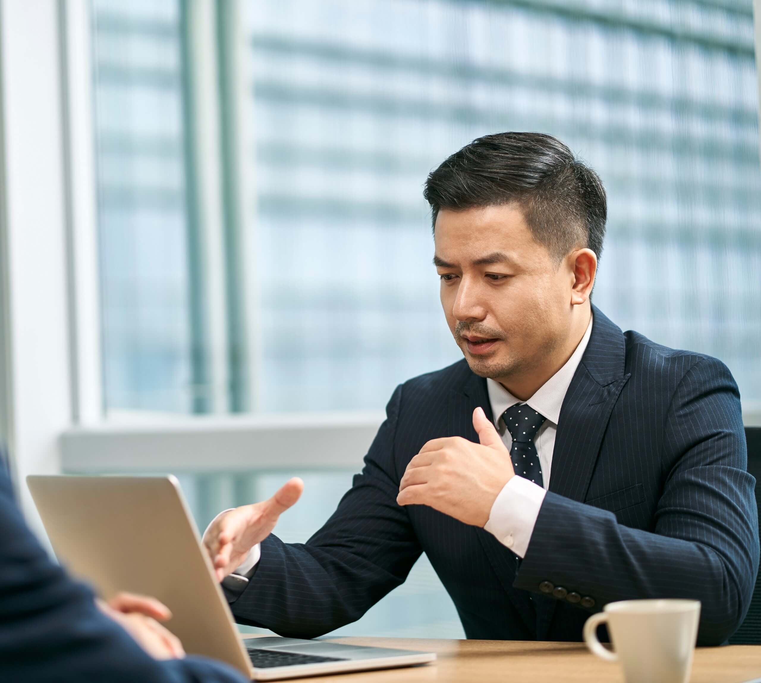 Two men in business suits sit at a table with a laptop and a coffee cup, having a serious conversation in a modern office with large windows.