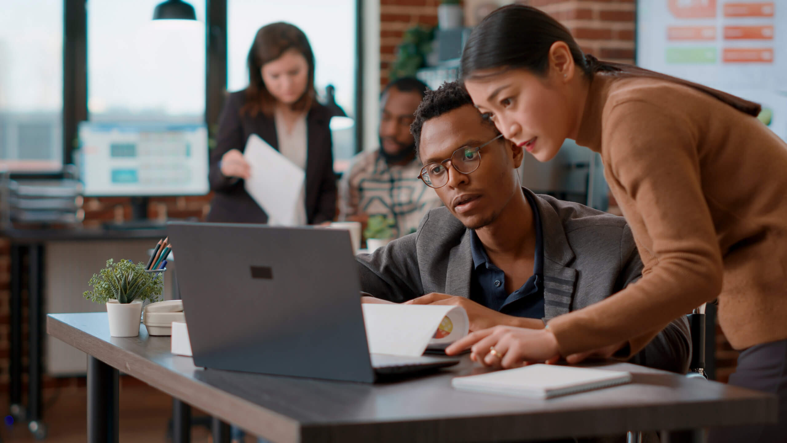 Two coworkers collaborate at a desk with a laptop and papers, while two other colleagues work in the background in a modern office setting.
