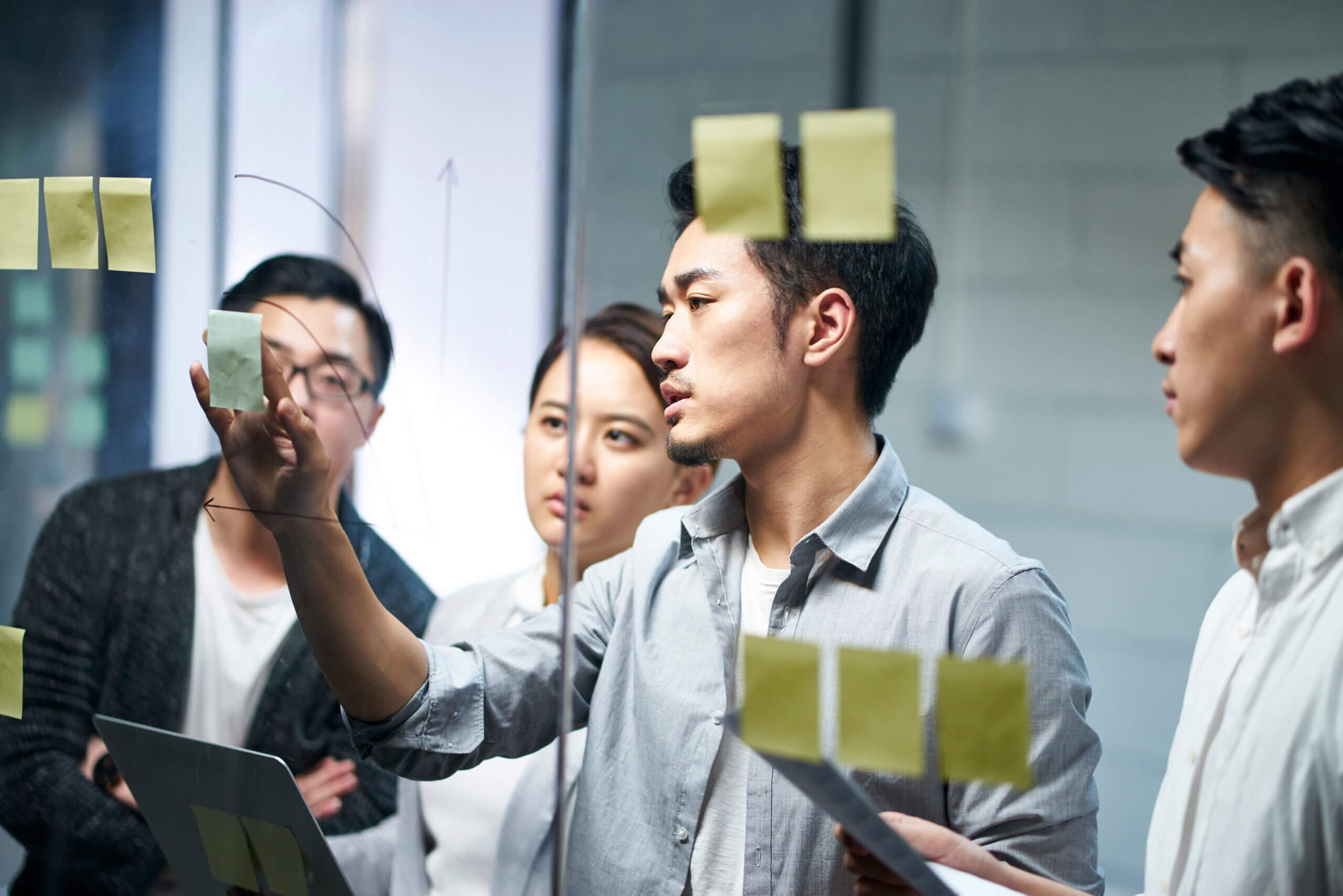 Four people collaborate in an office, discussing ideas and writing on a glass wall covered with sticky notes. One person points at the notes while others listen and observe attentively.