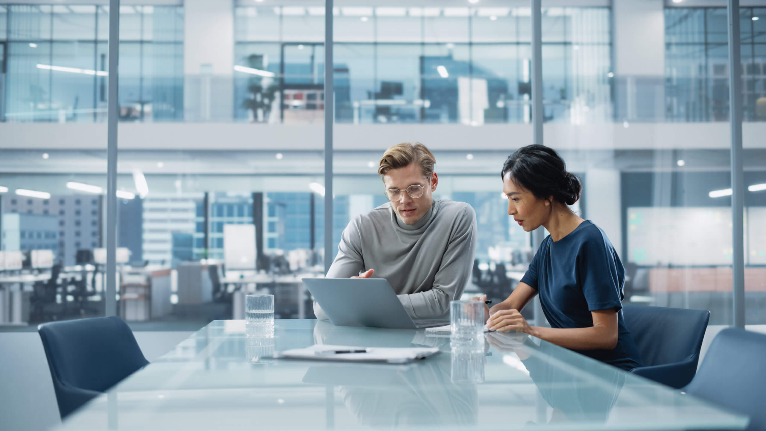 Two people sit at a glass conference table in a modern office, looking at a laptop. Papers and glasses of water are on the table. Large windows reveal more office space in the background.