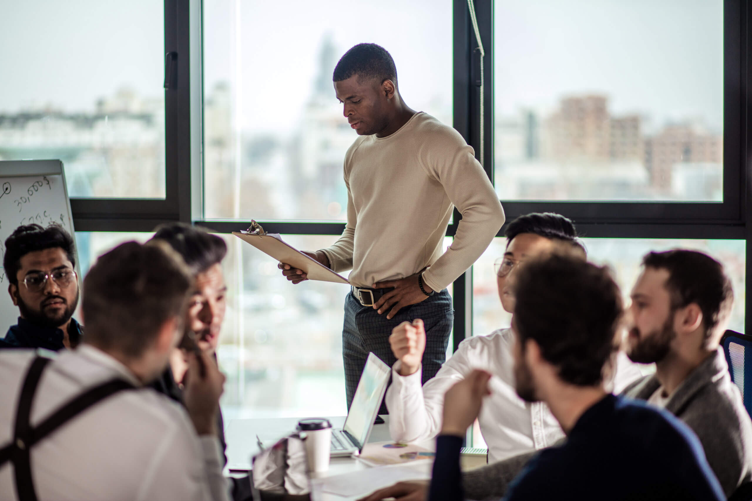 A man stands holding a clipboard while a group of people sit around a table in a modern office, engaged in discussion with laptops and papers. Large windows show a blurred cityscape in the background.