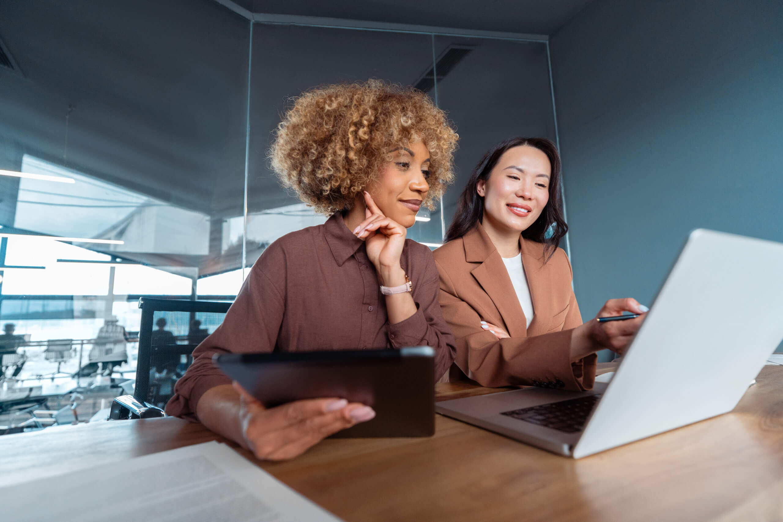 Two women sit at a desk in an office, one holding a tablet and the other pointing at a laptop screen. They appear to be having a discussion or working on a project together.