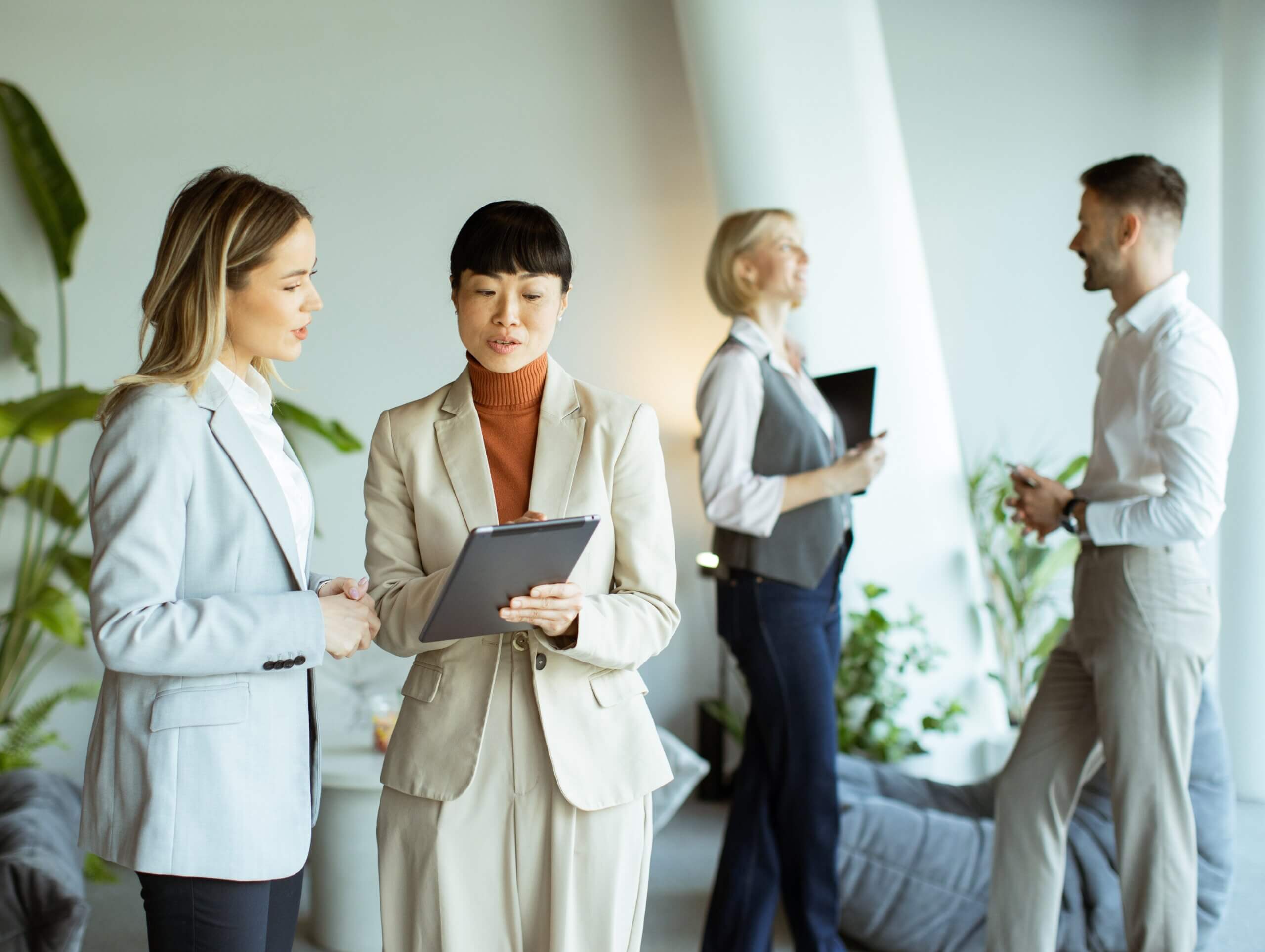 Two women in business attire discuss documents on a clipboard. In the background, a man and a woman talk, all standing in a bright office with plants and large windows.