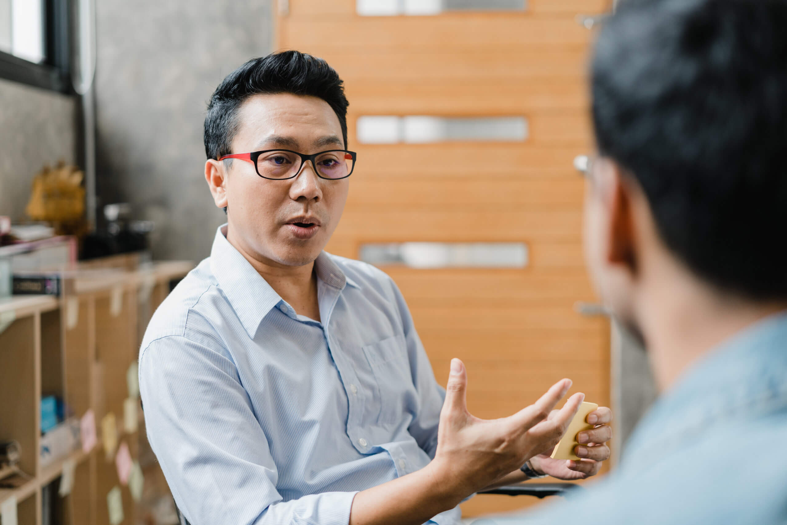 A man with glasses and a light blue shirt speaks seriously to another person in an indoor setting, gesturing with his hand.