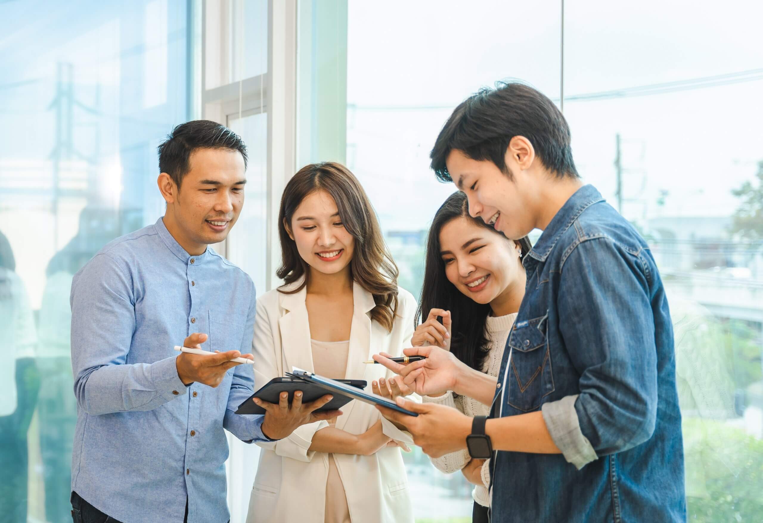 Four young adults stand together in a bright office, smiling as they review documents and take notes, suggesting a collaborative and positive work environment.