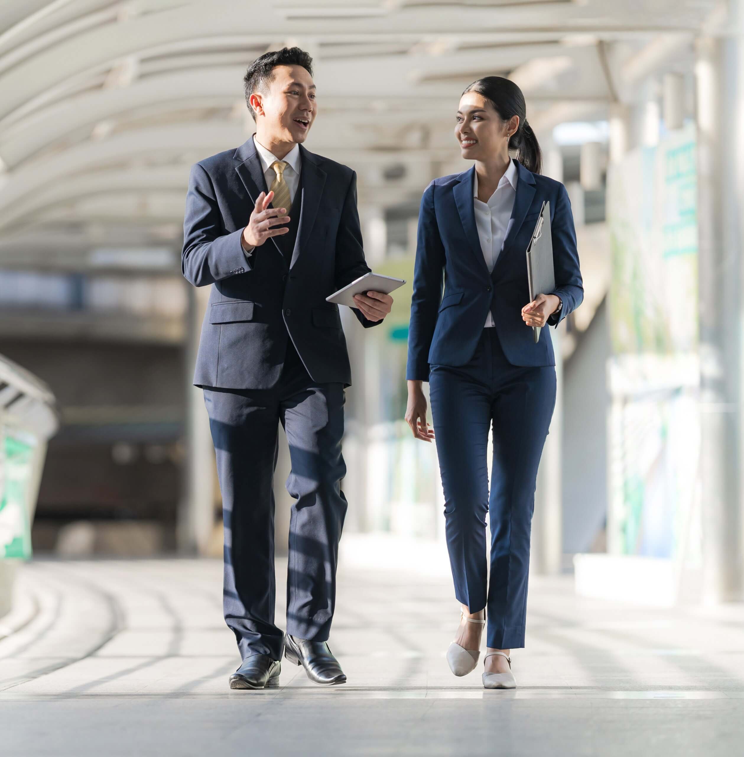 Two business professionals in suits walk side by side in a covered walkway, engaged in conversation. The man holds a tablet and gestures, while the woman carries a folder and smiles. The setting is bright and modern.