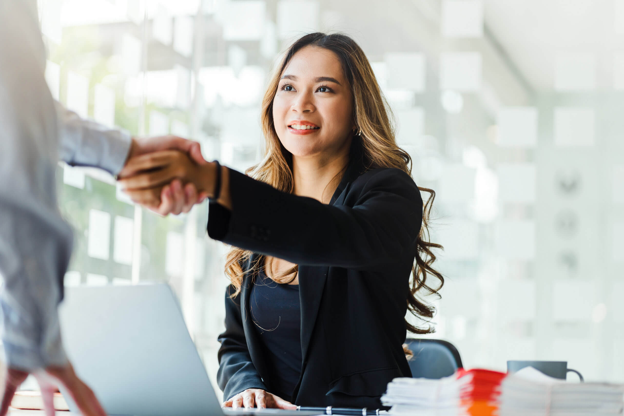 A woman in a black blazer smiles while shaking hands with someone across a desk in a bright, modern office setting. A laptop and neatly stacked papers are visible on the desk.