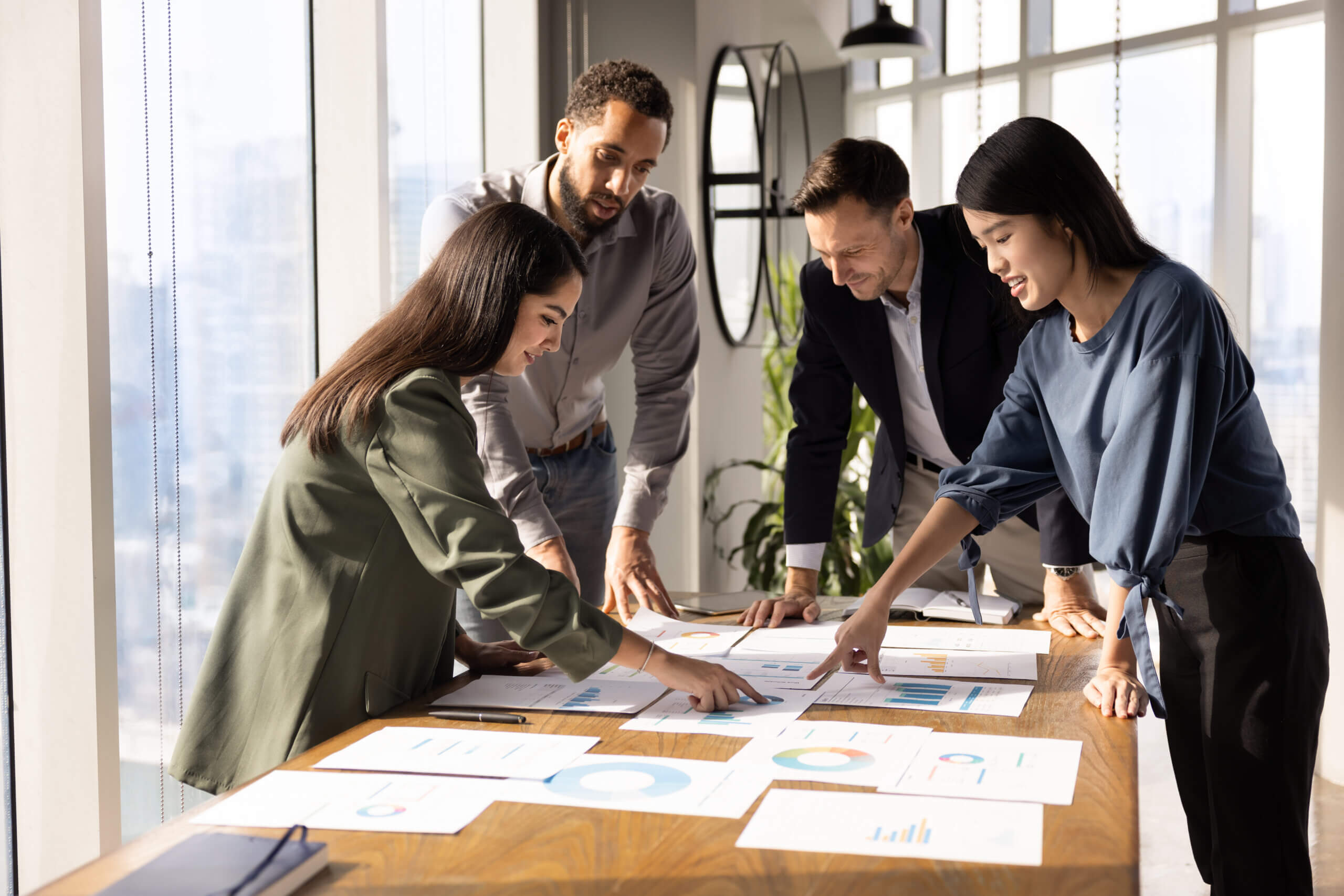 Four people stand around a table, discussing and pointing at charts and graphs on papers. They appear to be collaborating on a business project in a bright, modern office with large windows.