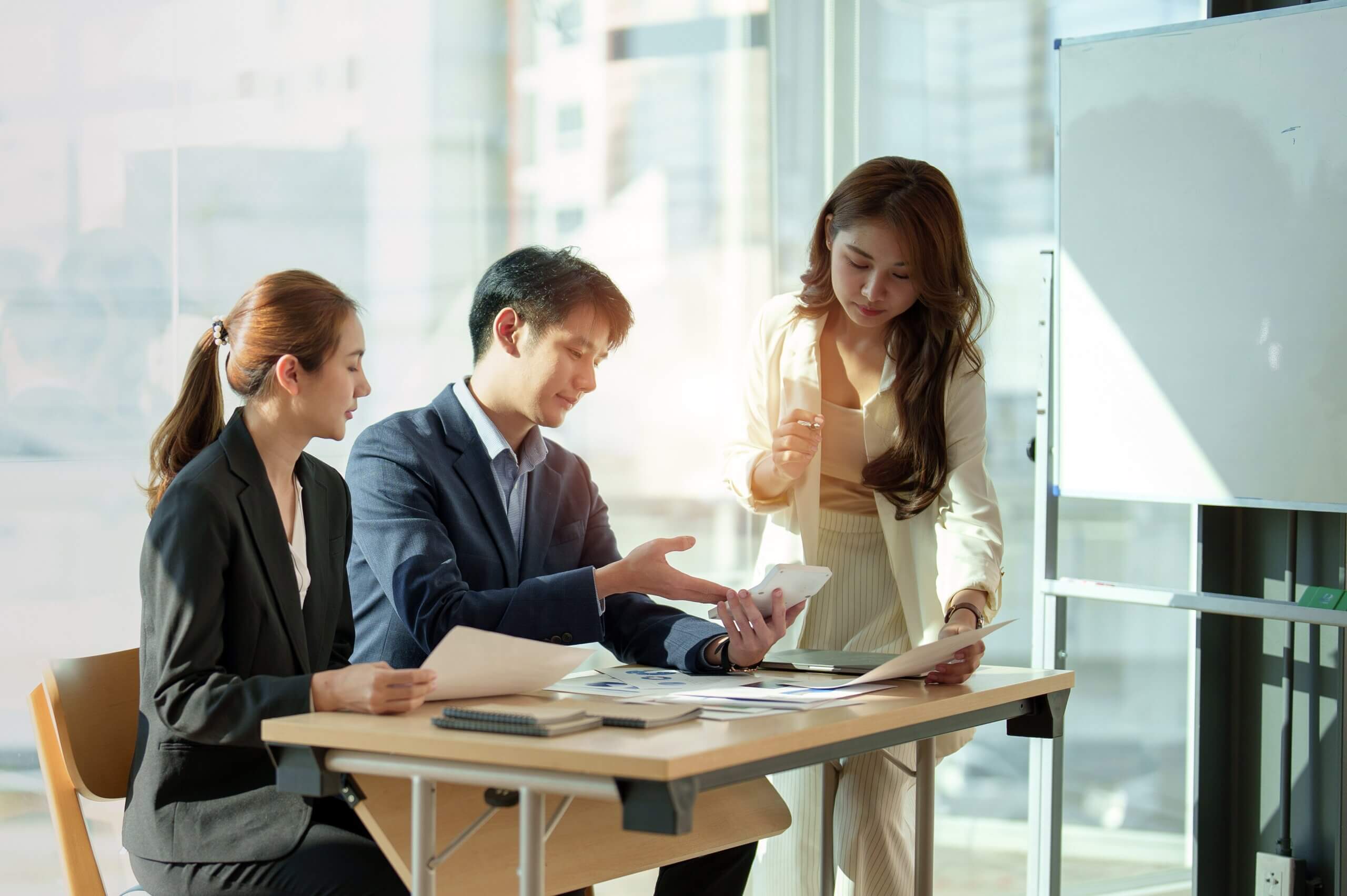 Three people in business attire sit and stand around a desk with papers, a calculator, and a tablet, discussing work in a brightly lit office with large windows and a whiteboard.