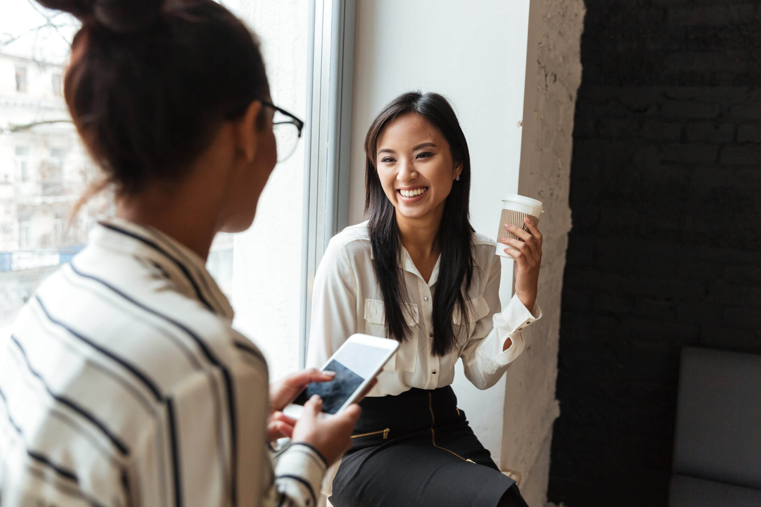 Two women are talking by a window; one is holding a phone and the other, smiling, holds a coffee cup. They appear to be having a friendly conversation in a bright, casual office setting.