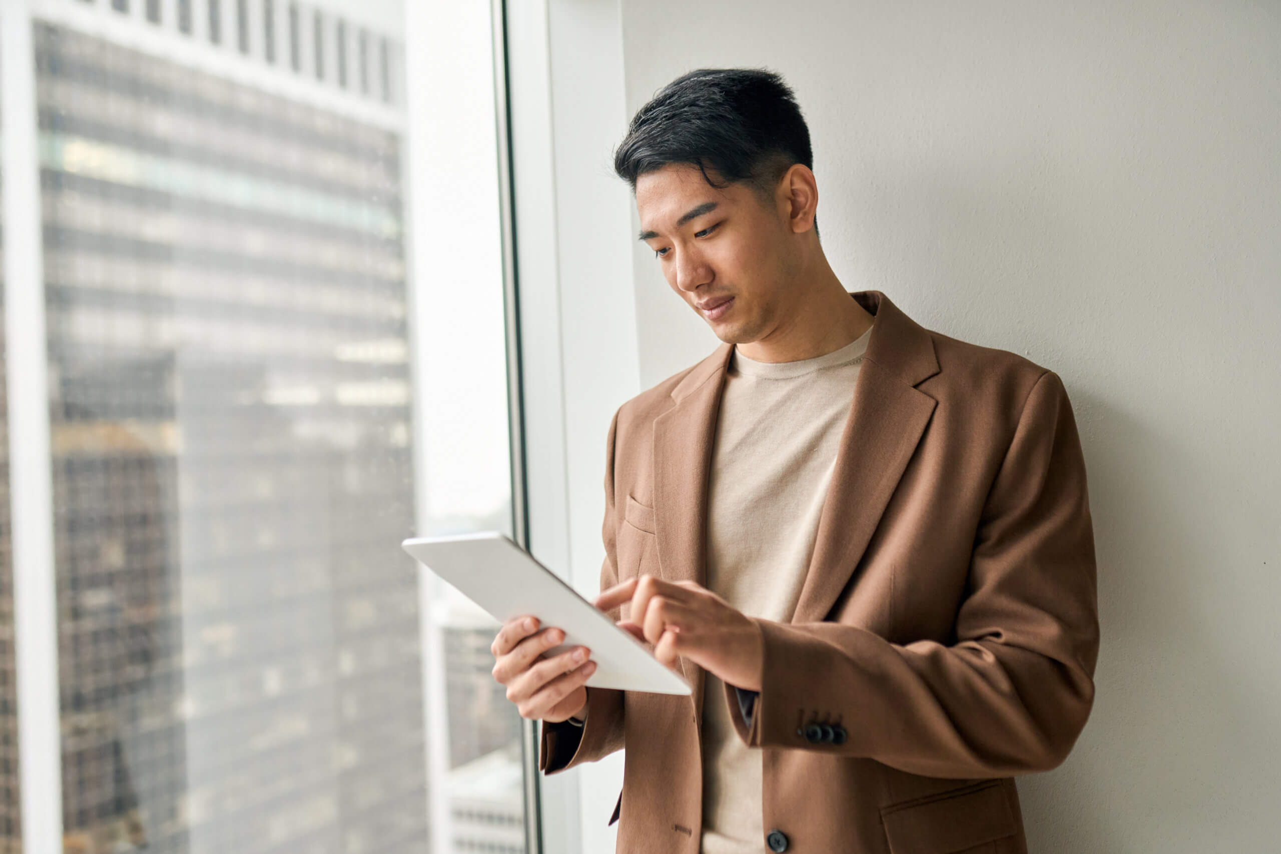 A man in a brown blazer stands by a window, using a tablet. A cityscape with tall buildings is visible in the background through the glass.