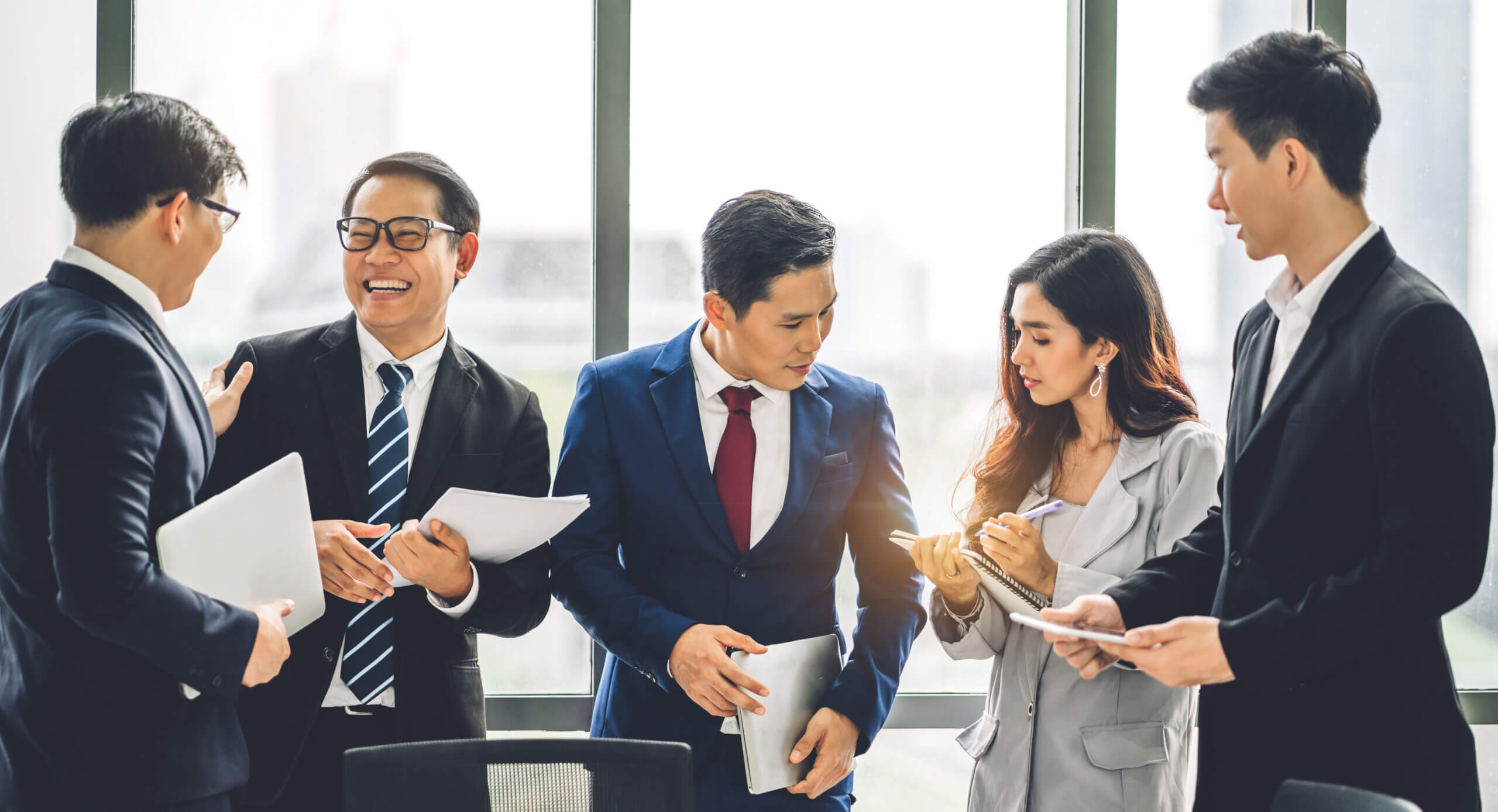 Five business professionals in formal attire are gathered in an office, talking and smiling while holding documents and tablets, with large windows in the background.