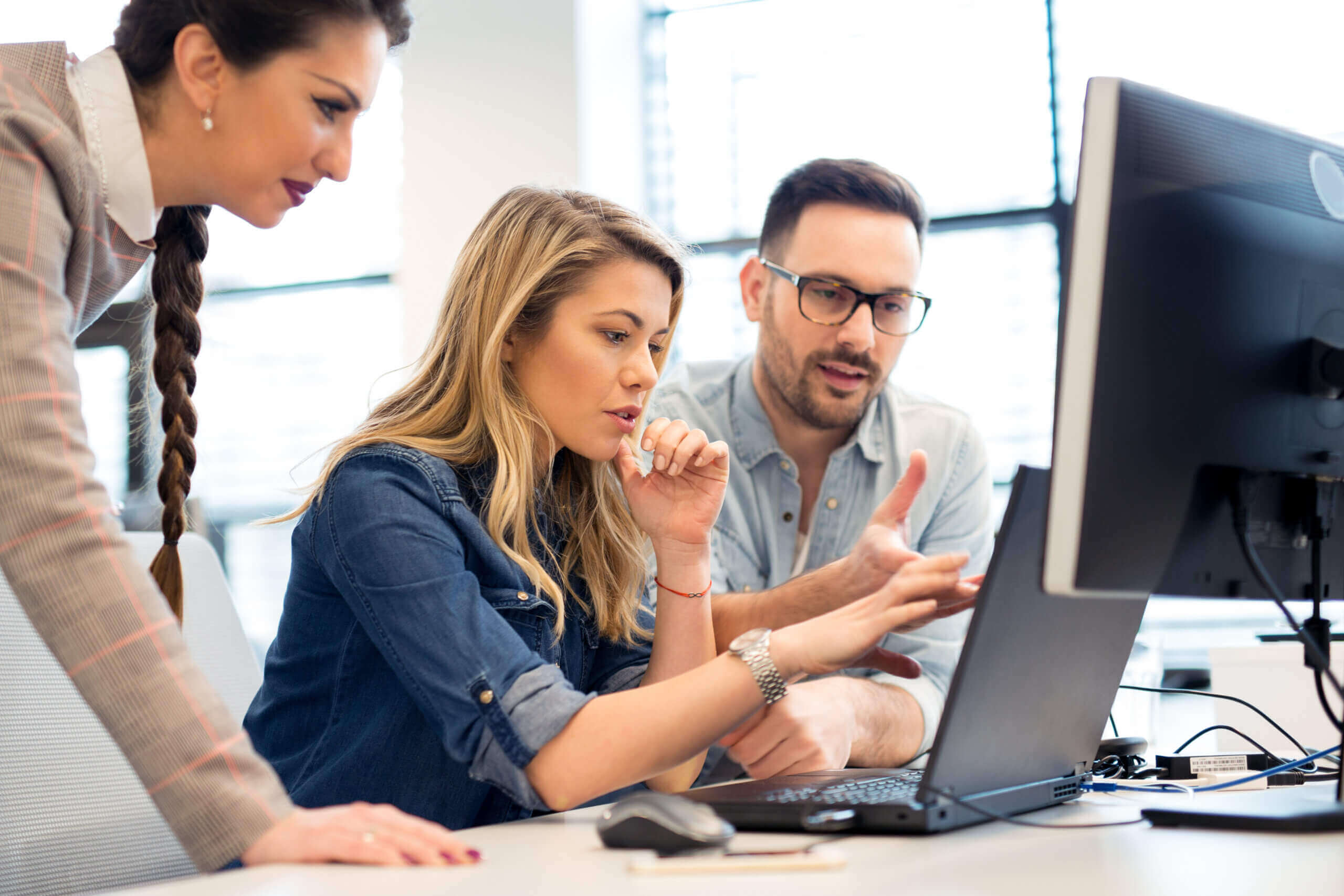 Three coworkers collaborate at a desk, looking intently at a computer monitor. One woman sits and gestures at the screen while the other two, a man and woman, stand close by, discussing and analyzing the content.