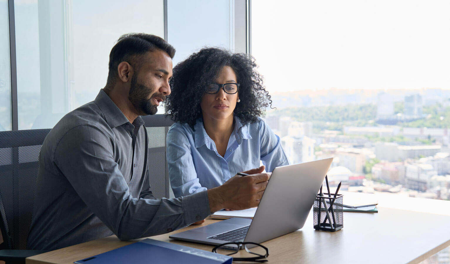 Two people sit at an office desk, looking at a laptop together. One person gestures toward the screen, while the other listens attentively. Large windows show a cityscape in the background.