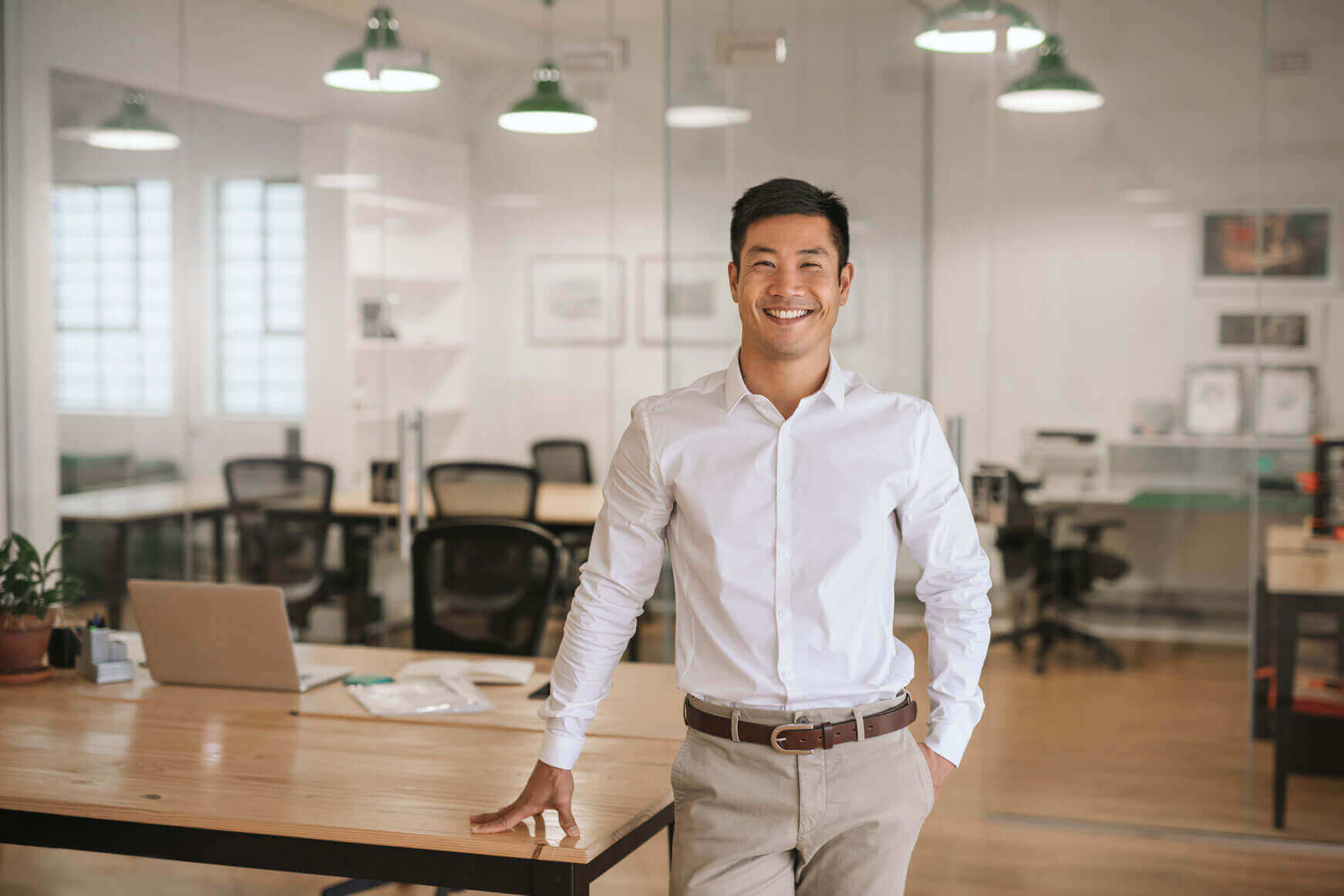 A man in a white shirt stands smiling in a modern office with glass walls, wooden desks, and empty chairs. A laptop and plant are on the desk beside him. The atmosphere is bright and professional.