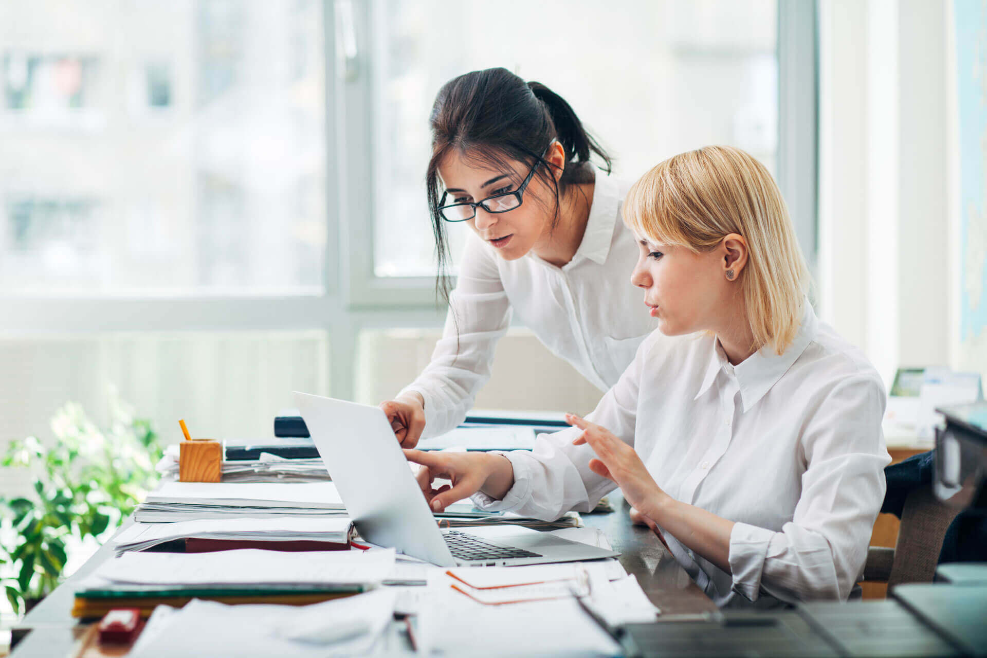 Two women in white shirts work together at a desk covered with papers, looking at a laptop screen. One is sitting and typing, while the other leans over, pointing at the laptop. Bright office setting with large windows.