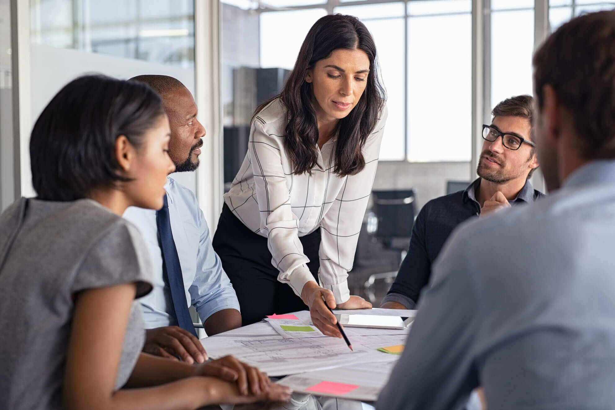 Five professionals in business attire sit around a table in a modern office. A woman stands, leaning forward to point at documents as the others listen attentively. Papers and sticky notes are spread on the table.