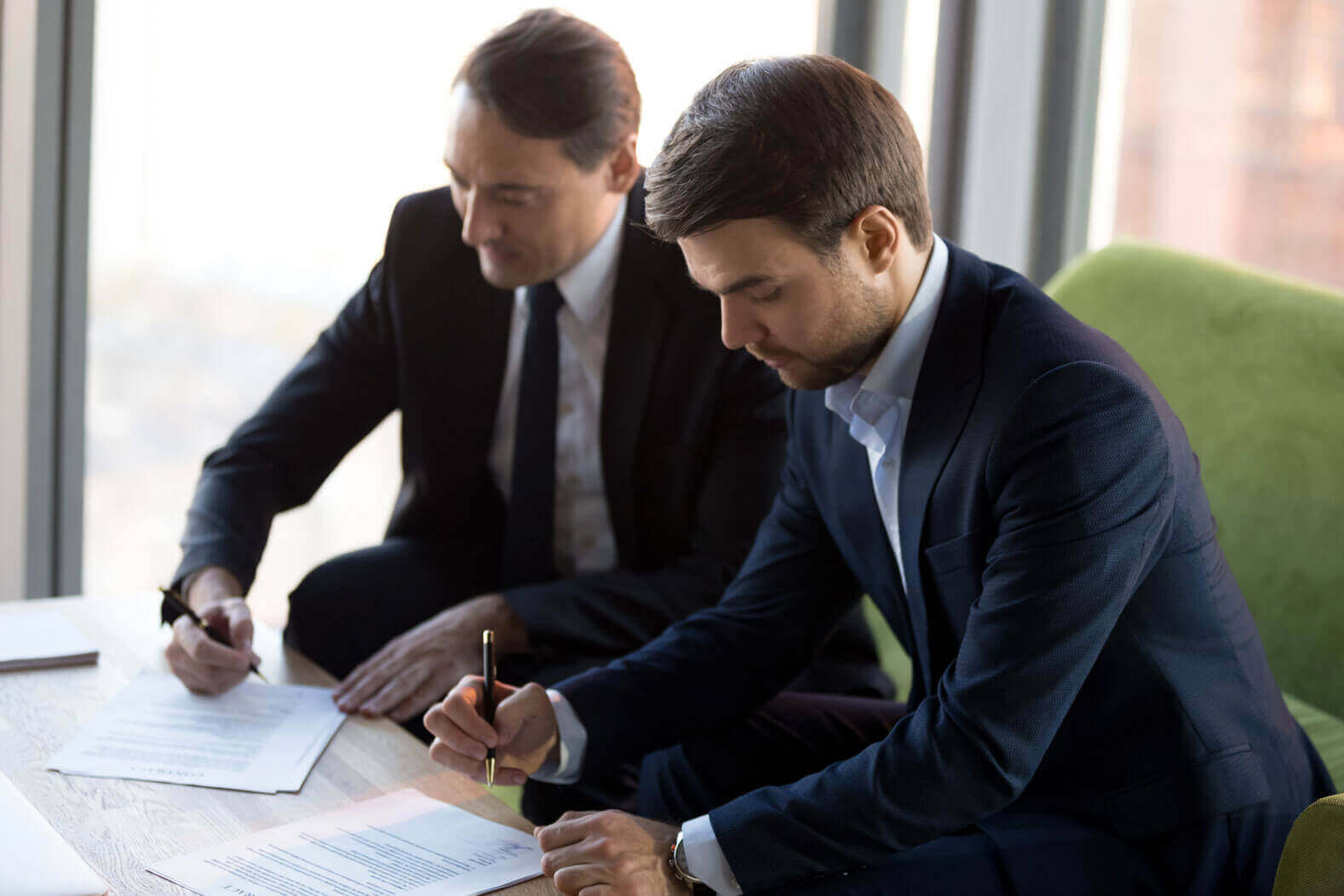 Two men in suits sit at a table, focused on signing documents with pens. Natural light comes through large windows behind them, creating a professional office setting.