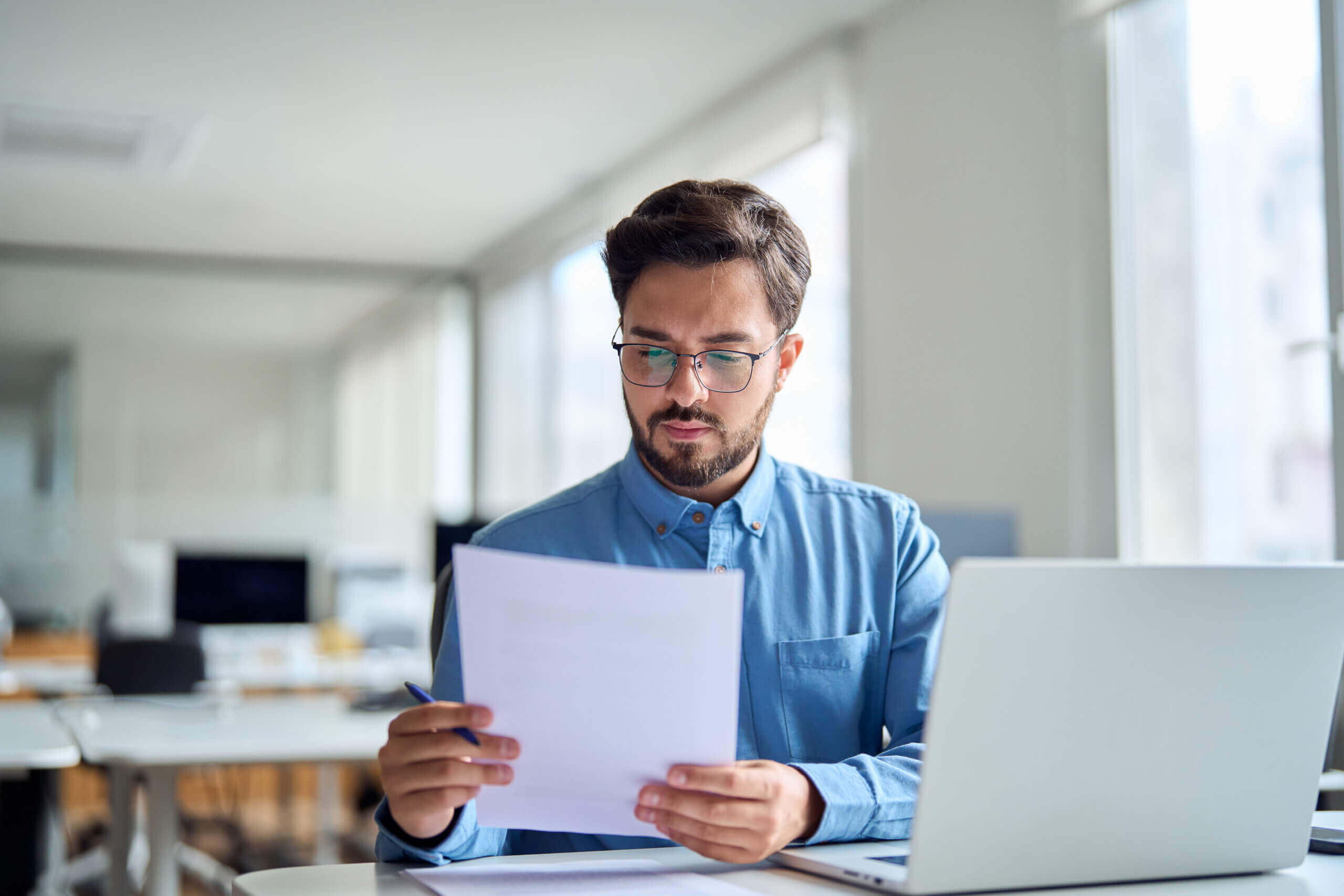 A man with glasses and a beard sits at a desk in an office, looking at documents and holding a pen, with a laptop open in front of him.