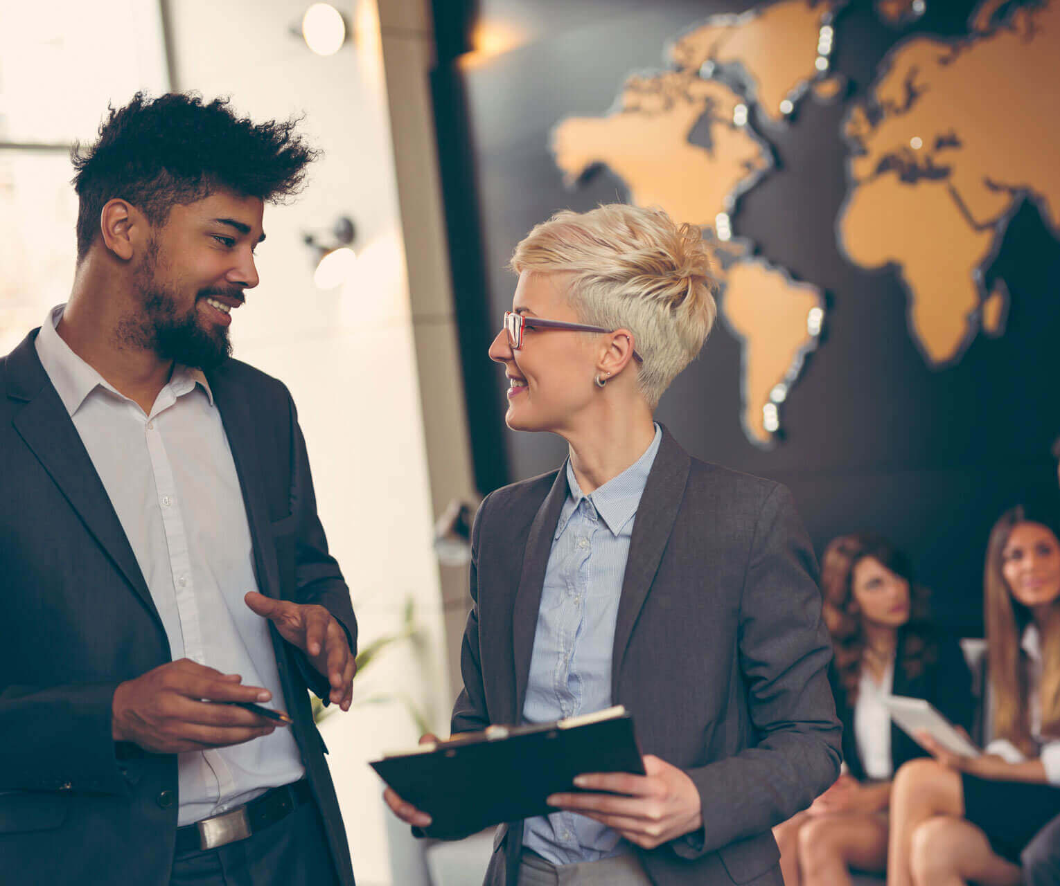 Two business professionals, a man and a woman, talk and smile in an office. The woman holds a clipboard. A world map is on the wall behind them, with several seated people in the background.
